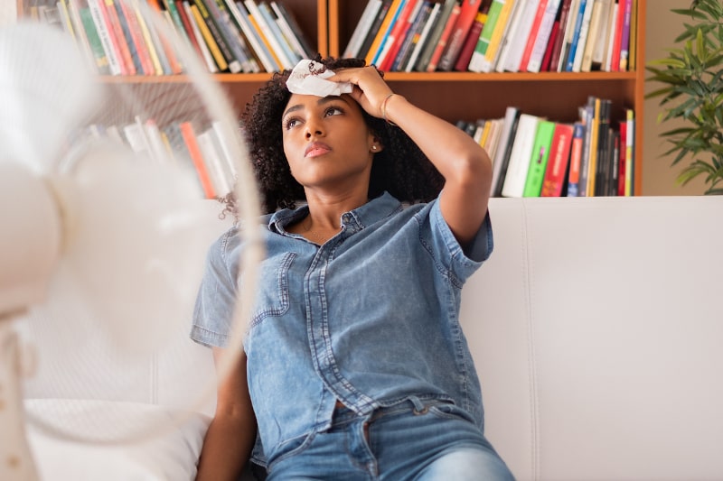 person sitting on couch in front of fan and dabbing sweat on forehead